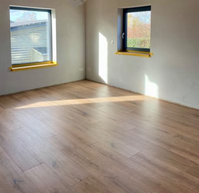 Heller Raum mit Holzfußboden und zwei Fenstern, die Sonnenlicht hereinlassen.