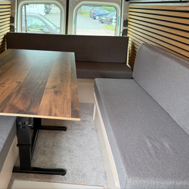 Kleine Wohnmobilausbau mit grauen Polstern und Tisch in einem Fahrzeug.