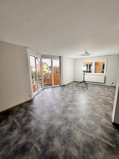 Heller, leerer Raum mit großem Fenster und Balkon, grauem Bodenbelag.